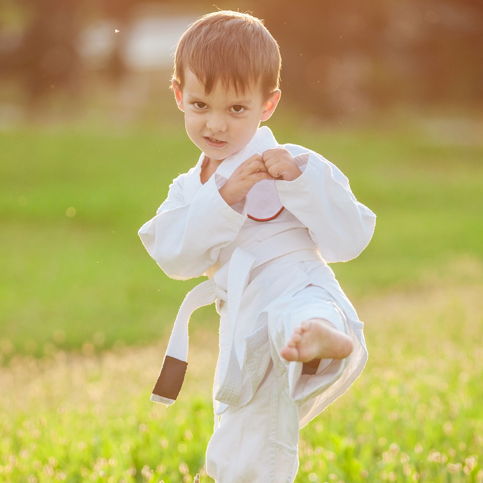 A young boy in a white karate uniform is standing on one leg in a field.