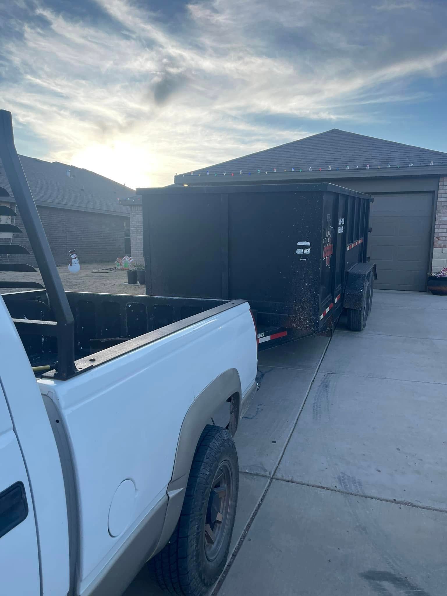 A white truck is towing a black dumpster in a driveway.