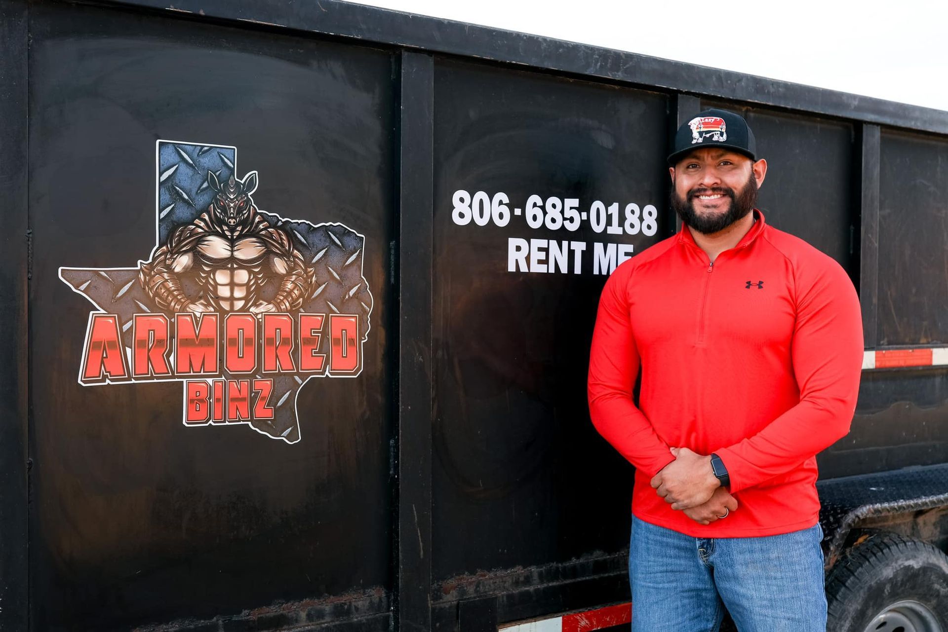 A man in a red shirt is standing in front of a dumpster.