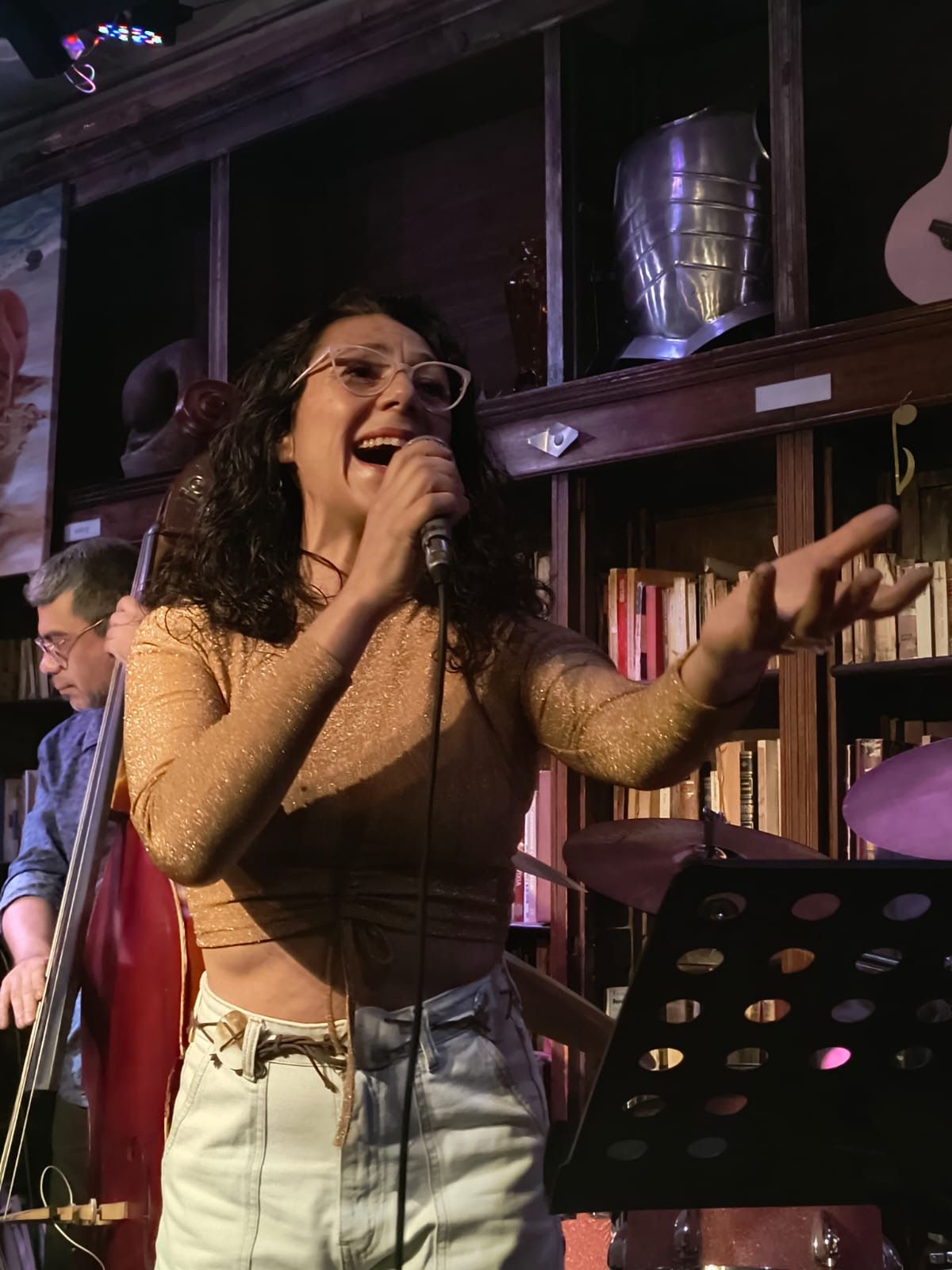 Mujer cantando con micrófono en el escenario; señala hacia su derecha, vestida con una blusa transparente y jeans.