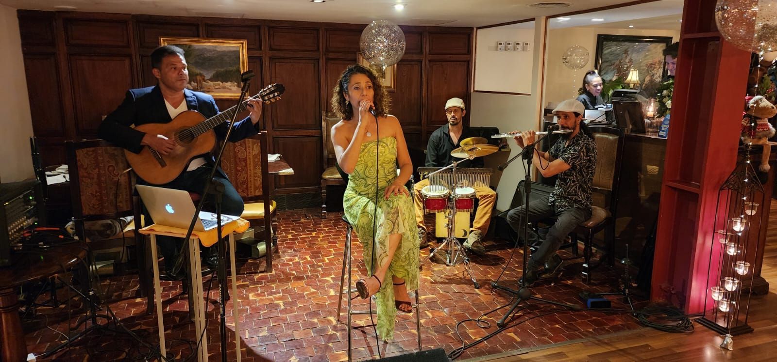 Una banda toca en vivo en una sala elegante. El guitarrista, el cantante y el baterista tocan, con confeti en el suelo.