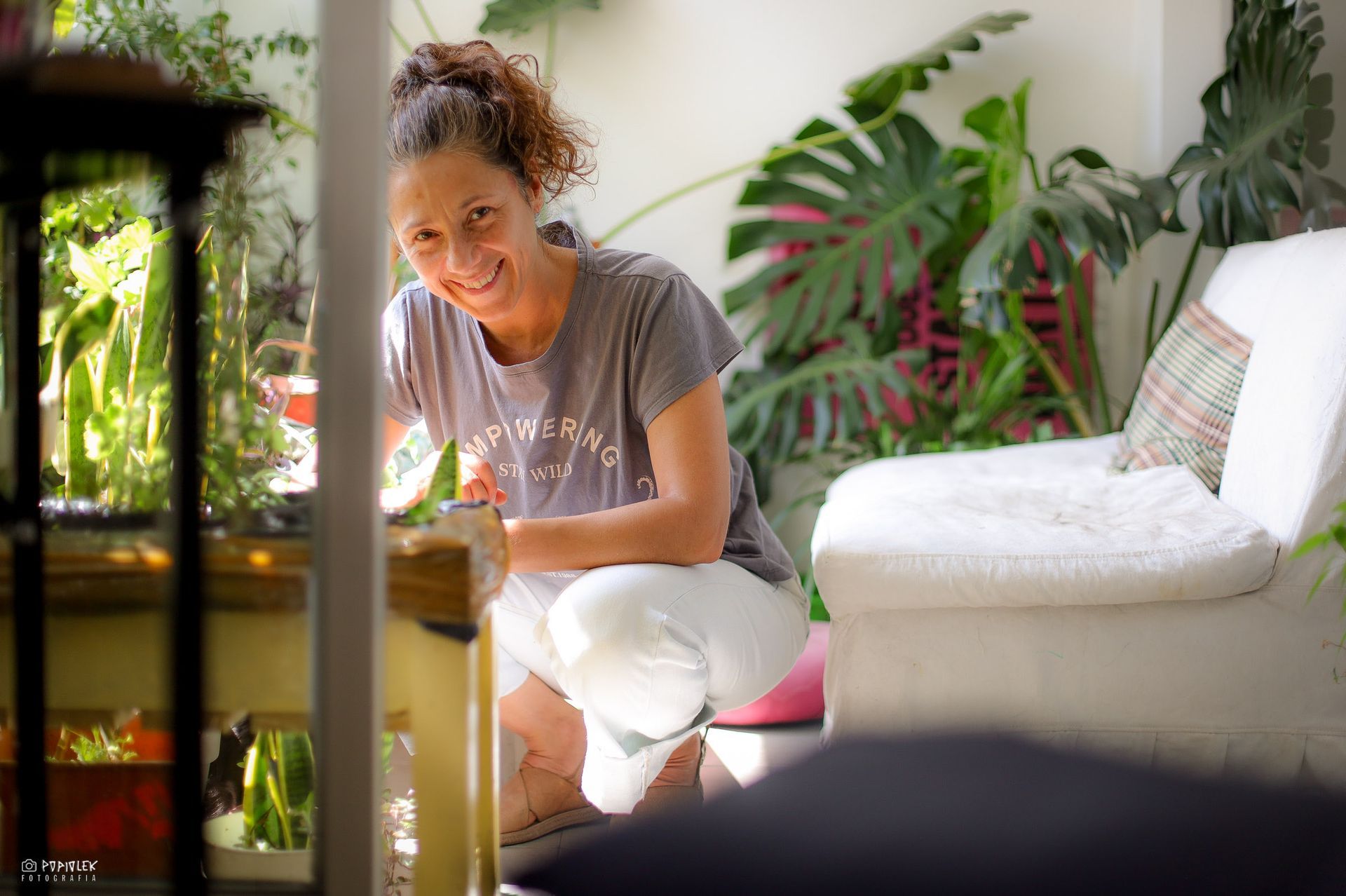 Una mujer con una camisa gris sonríe mientras cuida las plantas en un balcón soleado.
