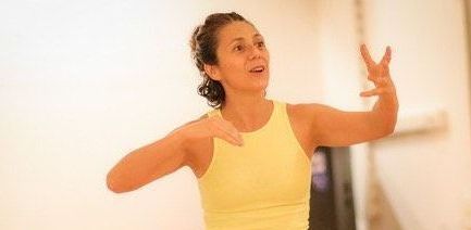 Mujer con camiseta amarilla gesticulando con las manos, posiblemente en un estudio de danza.