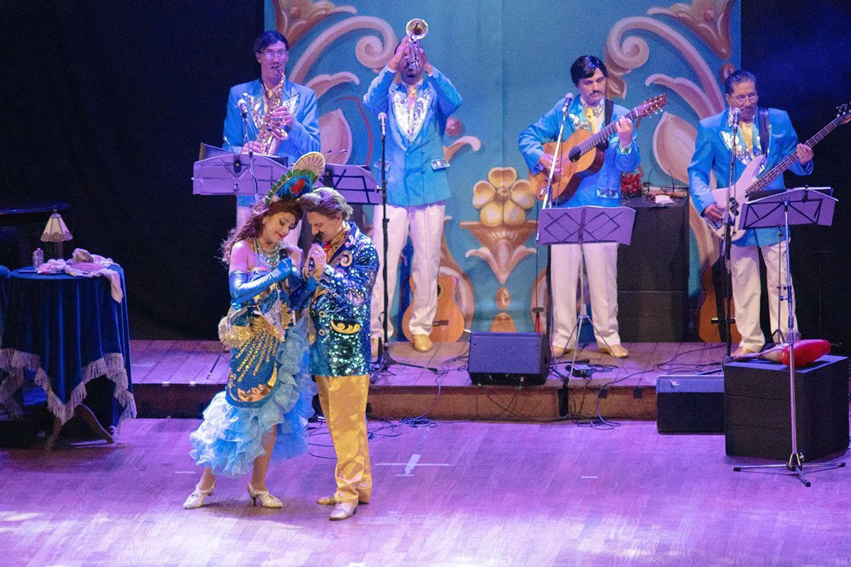 Una banda con trajes azules toca en el escenario; una pareja canta en el centro del escenario