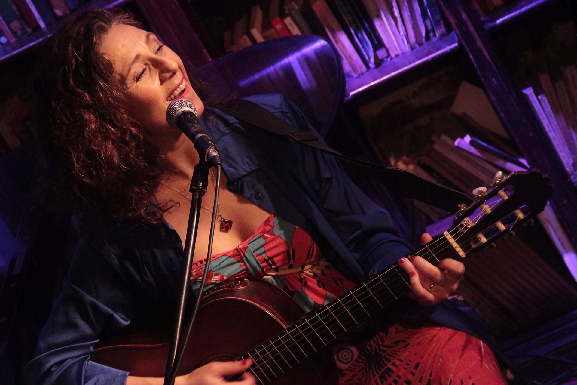 Mujer cantando y tocando la guitarra, con el escenario iluminado de púrpura. Estanterías de libros al fondo.