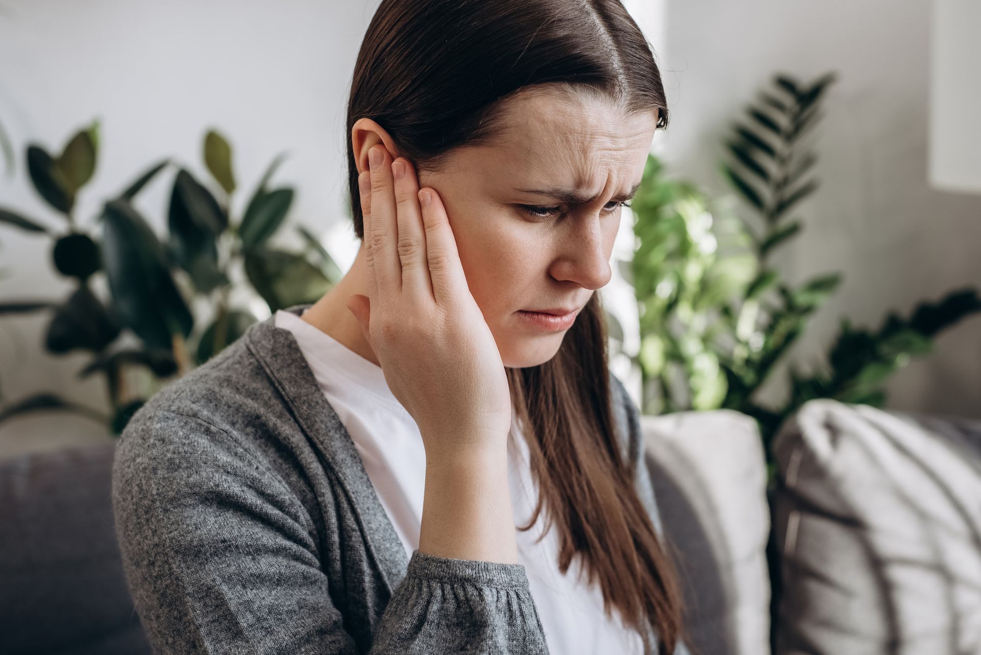 Woman holding her ear, grimacing, likely in pain. Indoors, near plants.