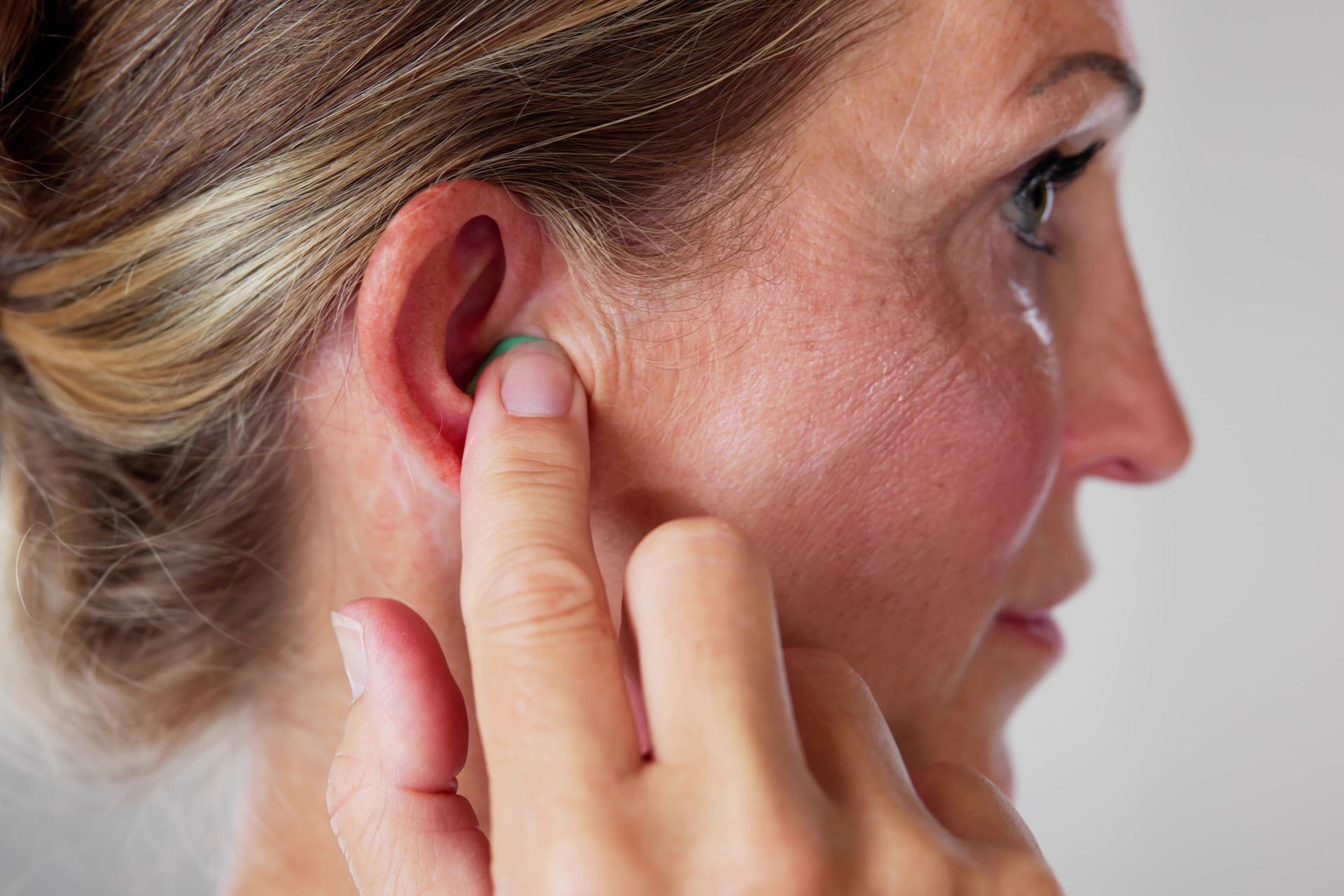 Woman with blonde hair inserts a green earplug into her ear with her finger.