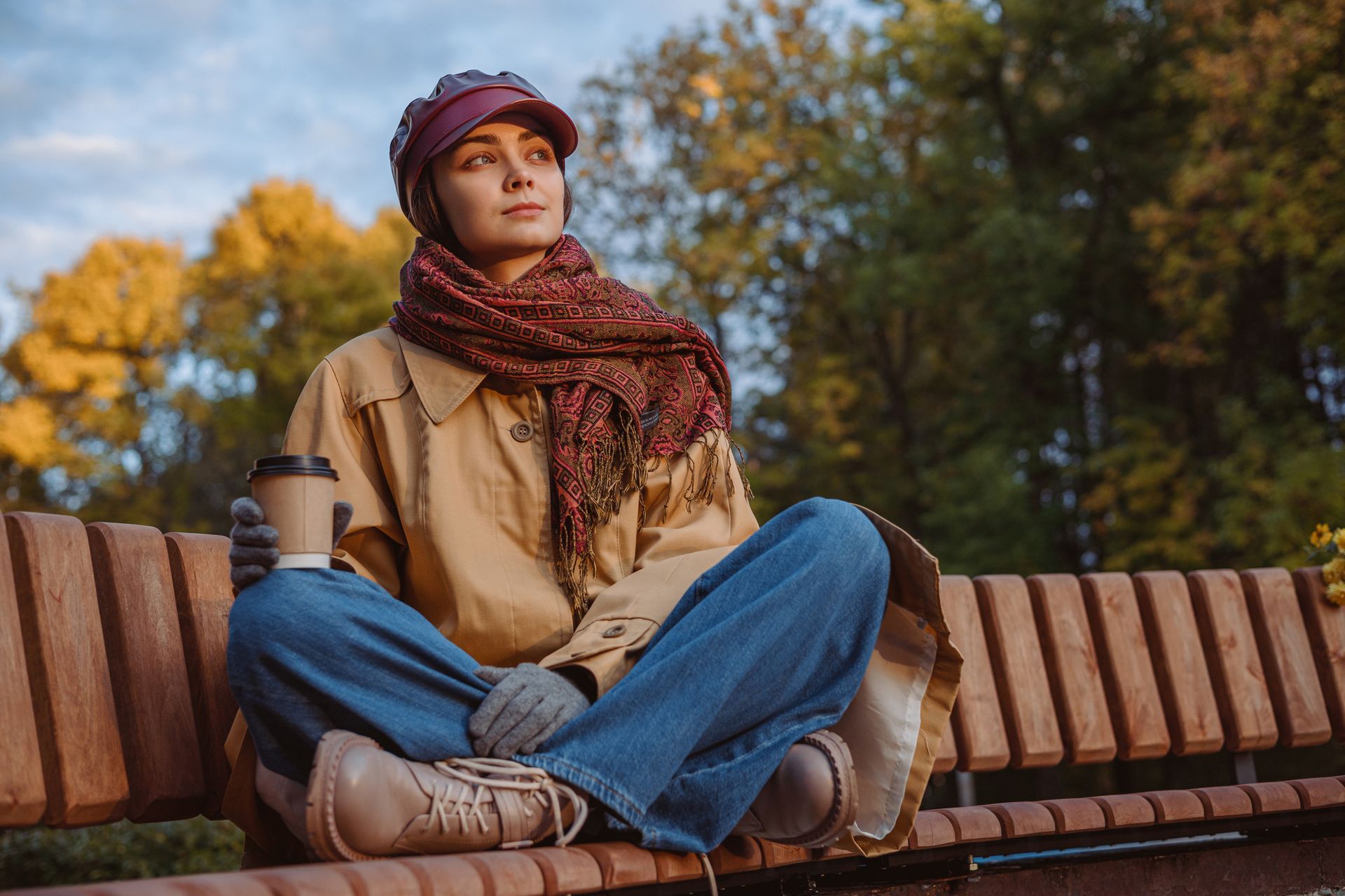 Woman in hat, scarf, coat, jeans, and gloves sits on a park bench with a coffee cup, looking upwards.