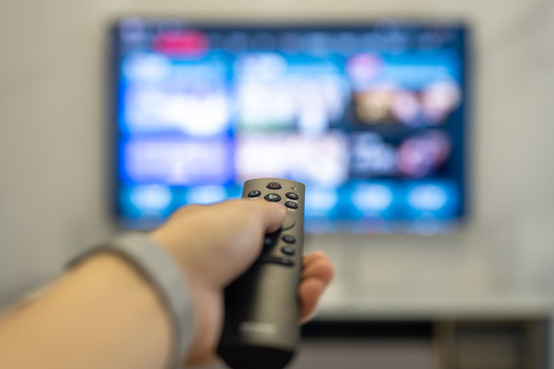 Hand holding a black remote, pointing at a blurred television displaying various options.