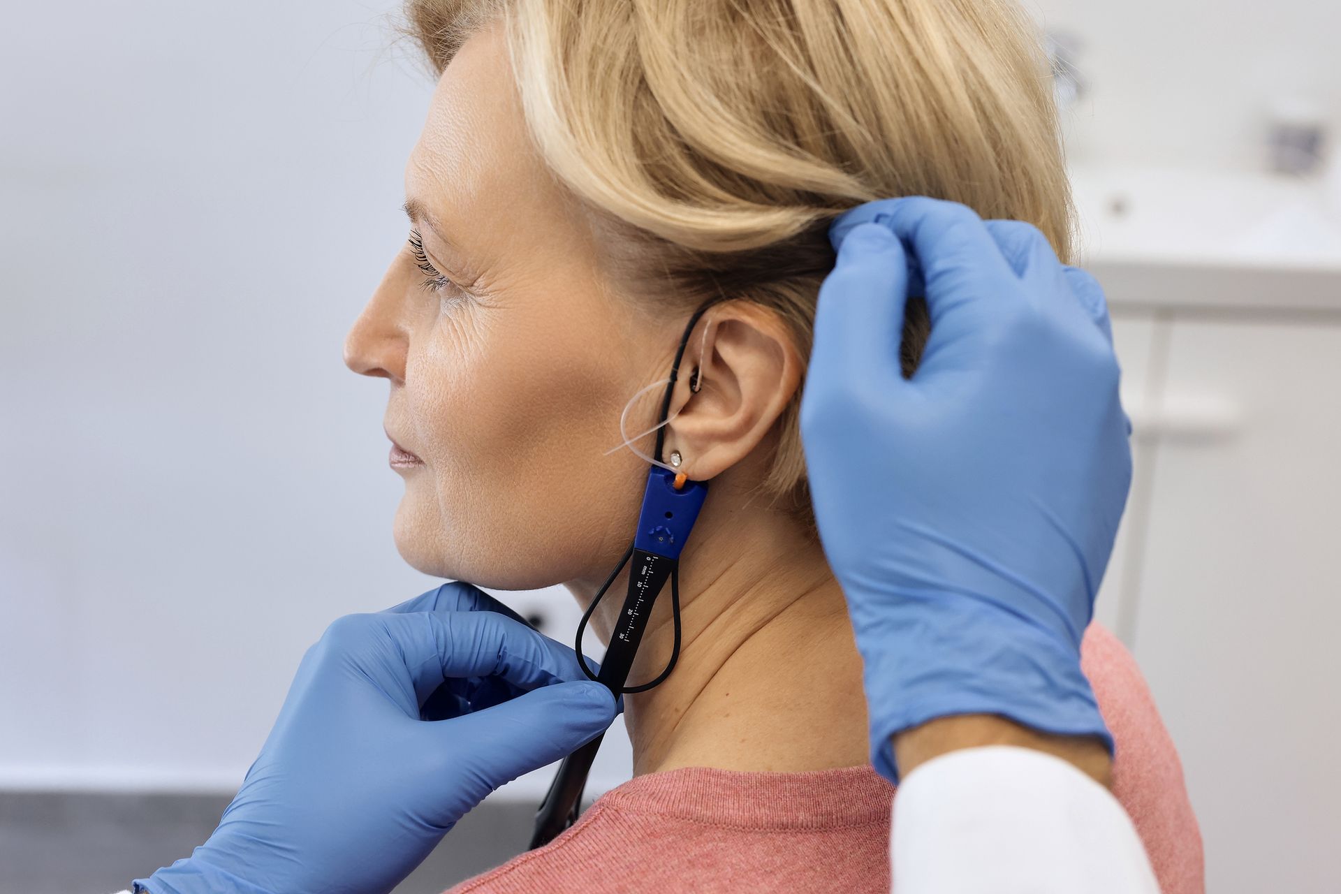 Person getting hearing test, earphone insertion. Doctor’s gloved hands, blonde woman, white room.