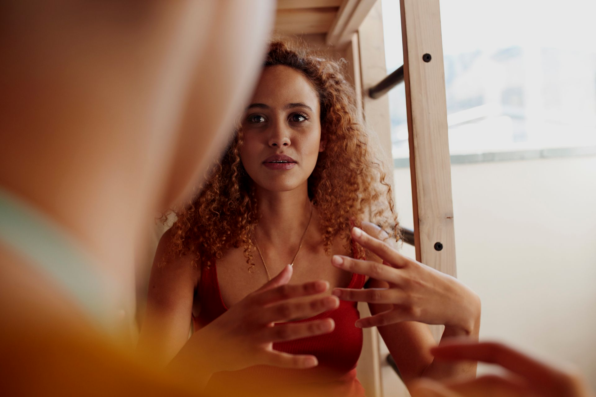 Woman with curly hair gesturing, talking to someone. Red top, wooden ladder, bright setting.