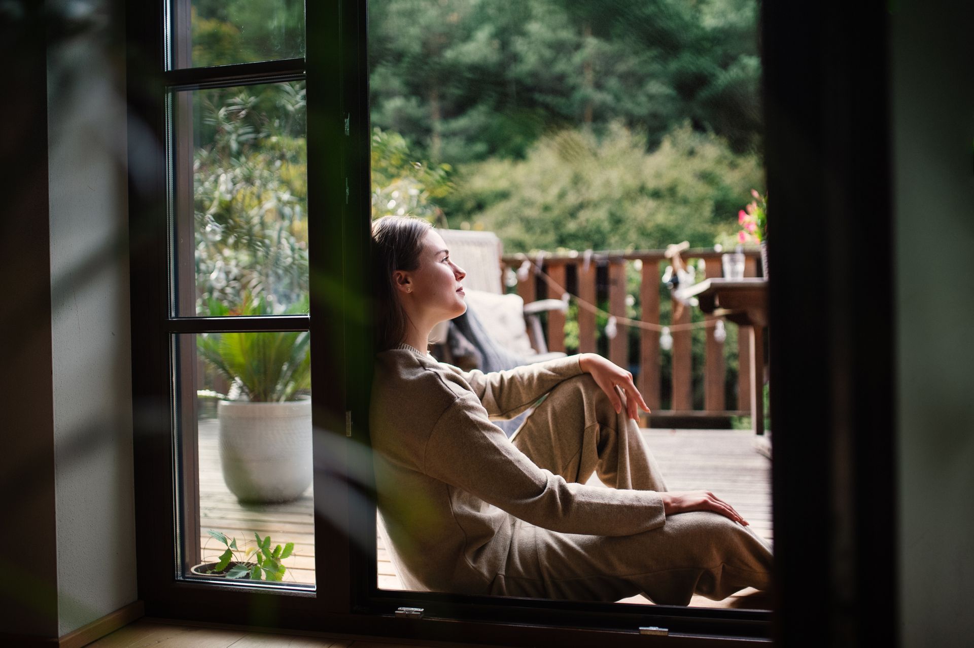 Woman in beige loungewear, sitting by open door, gazing at a sunlit wooden deck with green foliage in the background.