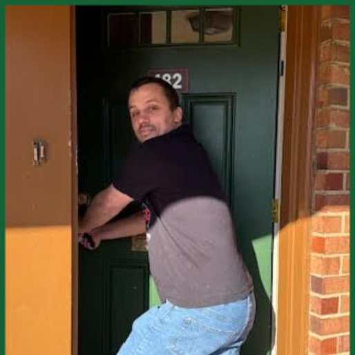 Man in blue jeans entering a green door, leaning forward, looking toward the camera. Exterior setting.
