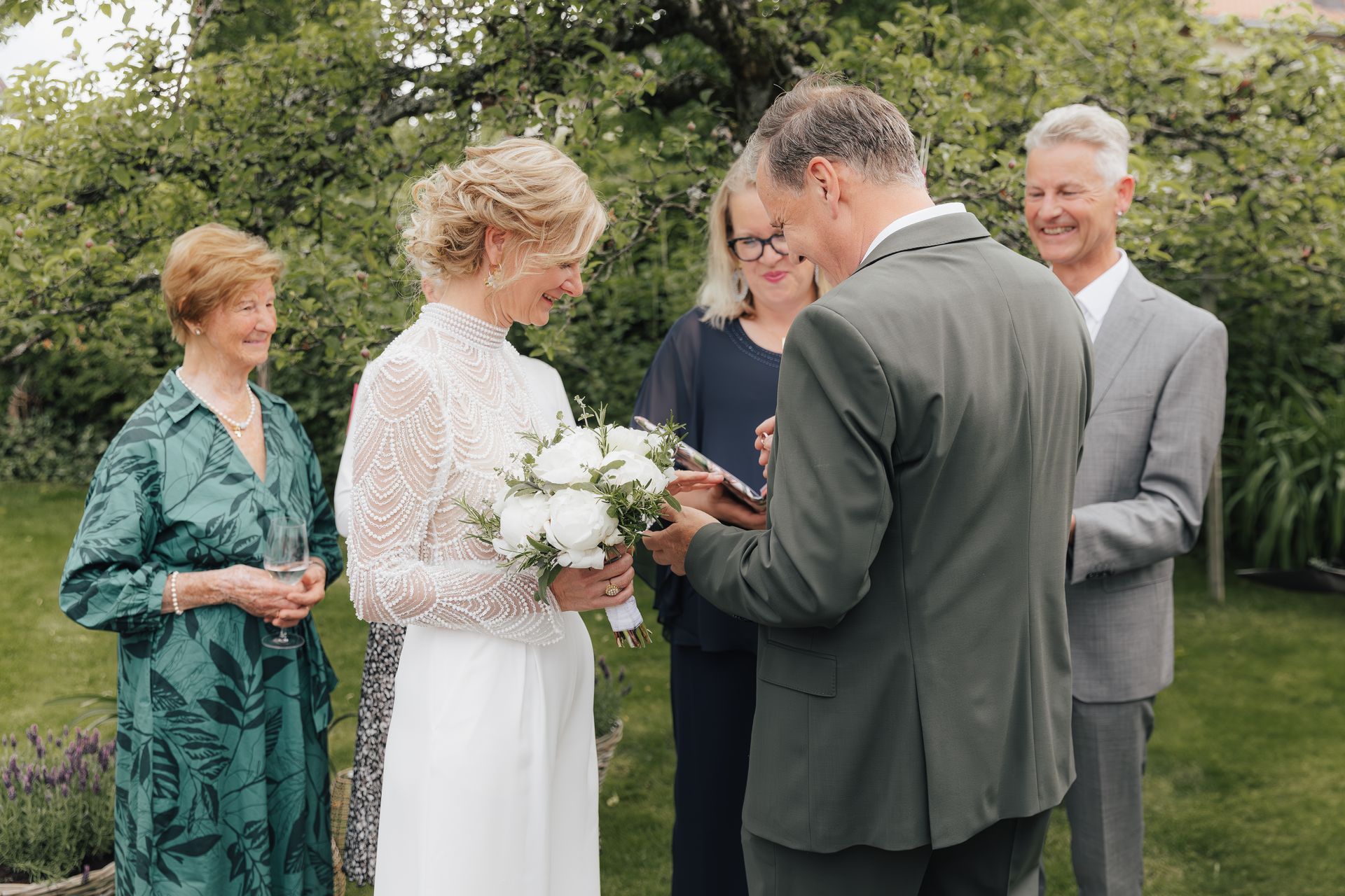 Hochzeitszeremonie: Braut im weißen Kleid mit Blumen, Bräutigam im grünen Anzug, Geistlicher, Gäste im Freien.