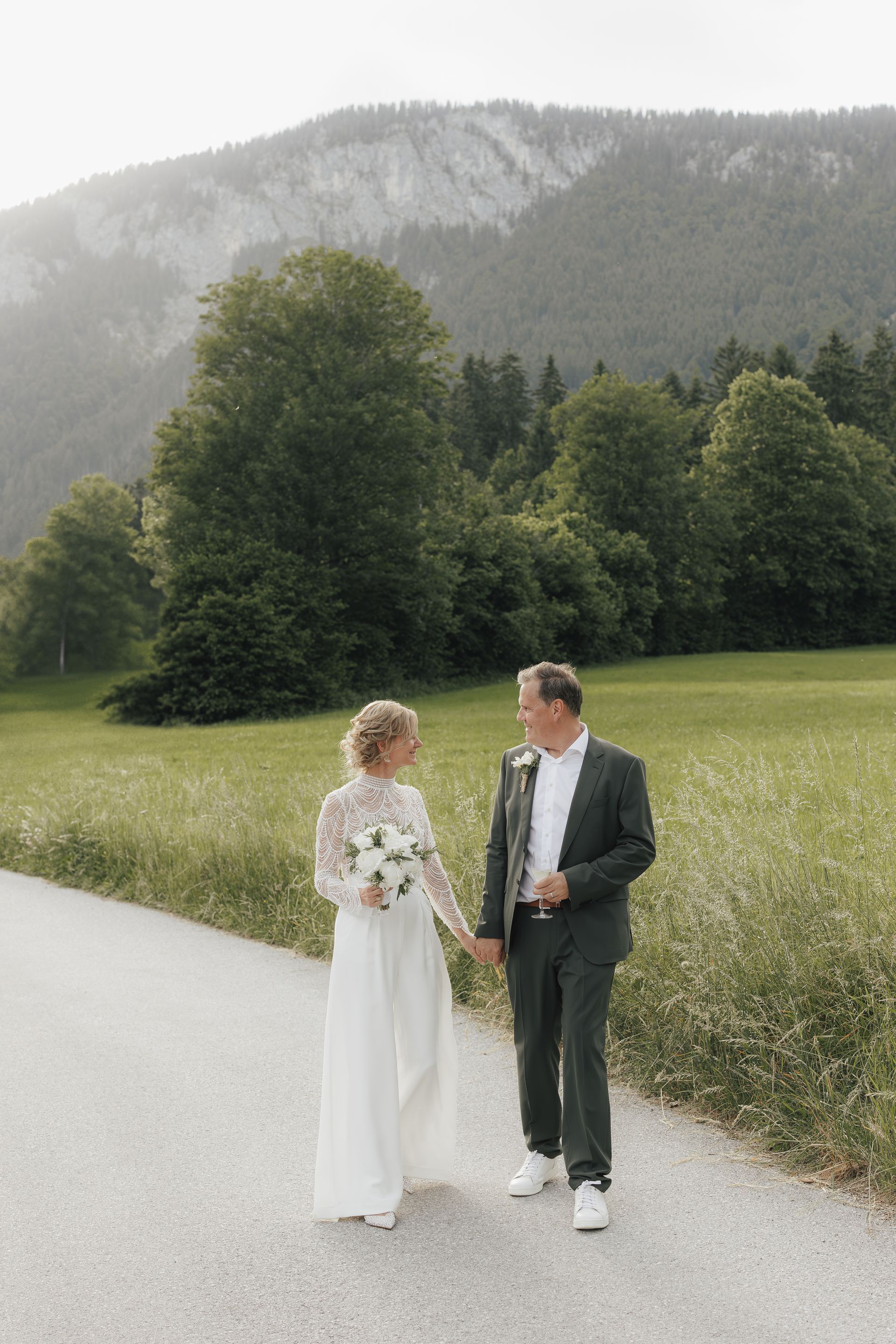 Paar in Hochzeitskleidung geht Händchen haltend auf einem Pfad mit Bergkulisse.