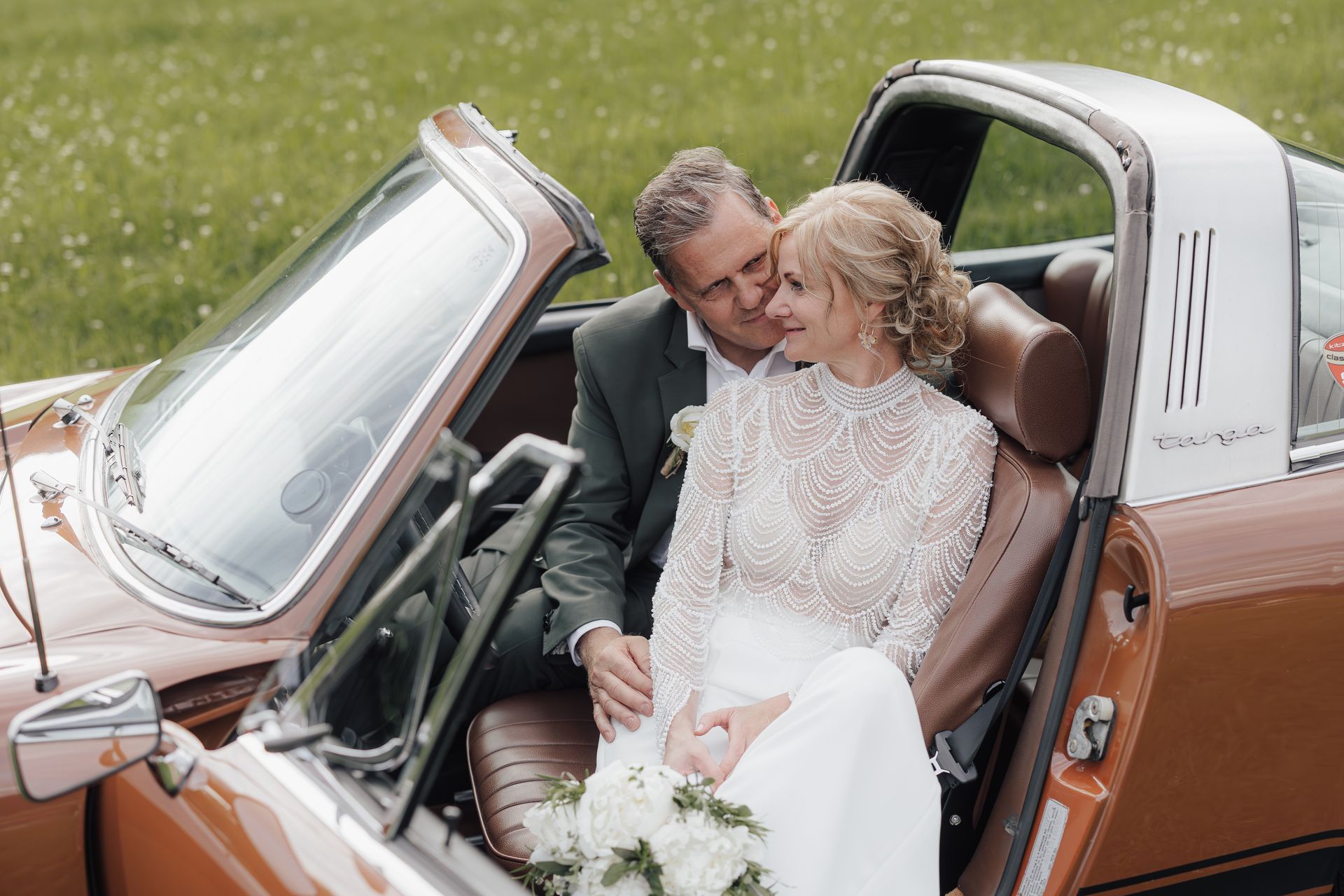 Frischvermählte sitzen in einem orangefarbenen Oldtimer-Sportwagen und schauen sich an