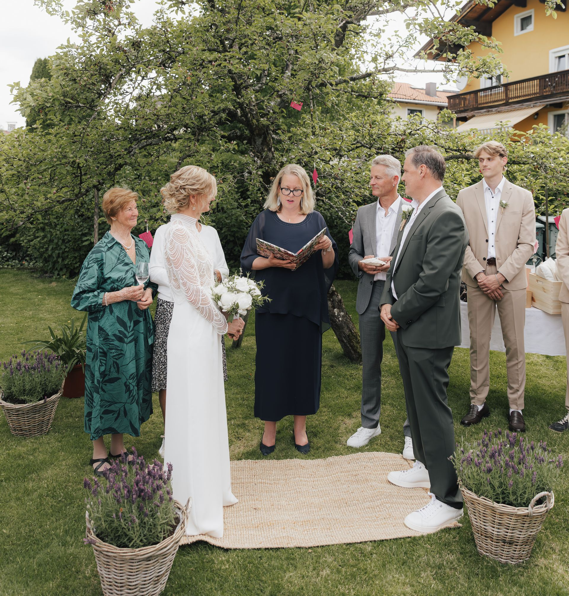 Hochzeitszeremonie im Freien mit Braut, Bräutigam, Geistlichem und Gästen in einer Gartenumgebung.
