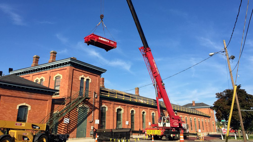 A red car is being lifted by a crane in front of a brick building.