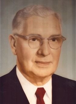 Portrait of an elderly man in a dark suit, glasses, and red tie. He has short, grey hair.