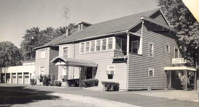 Black and white photo of a two-story building with porch, awning, and garage on a sunny day.