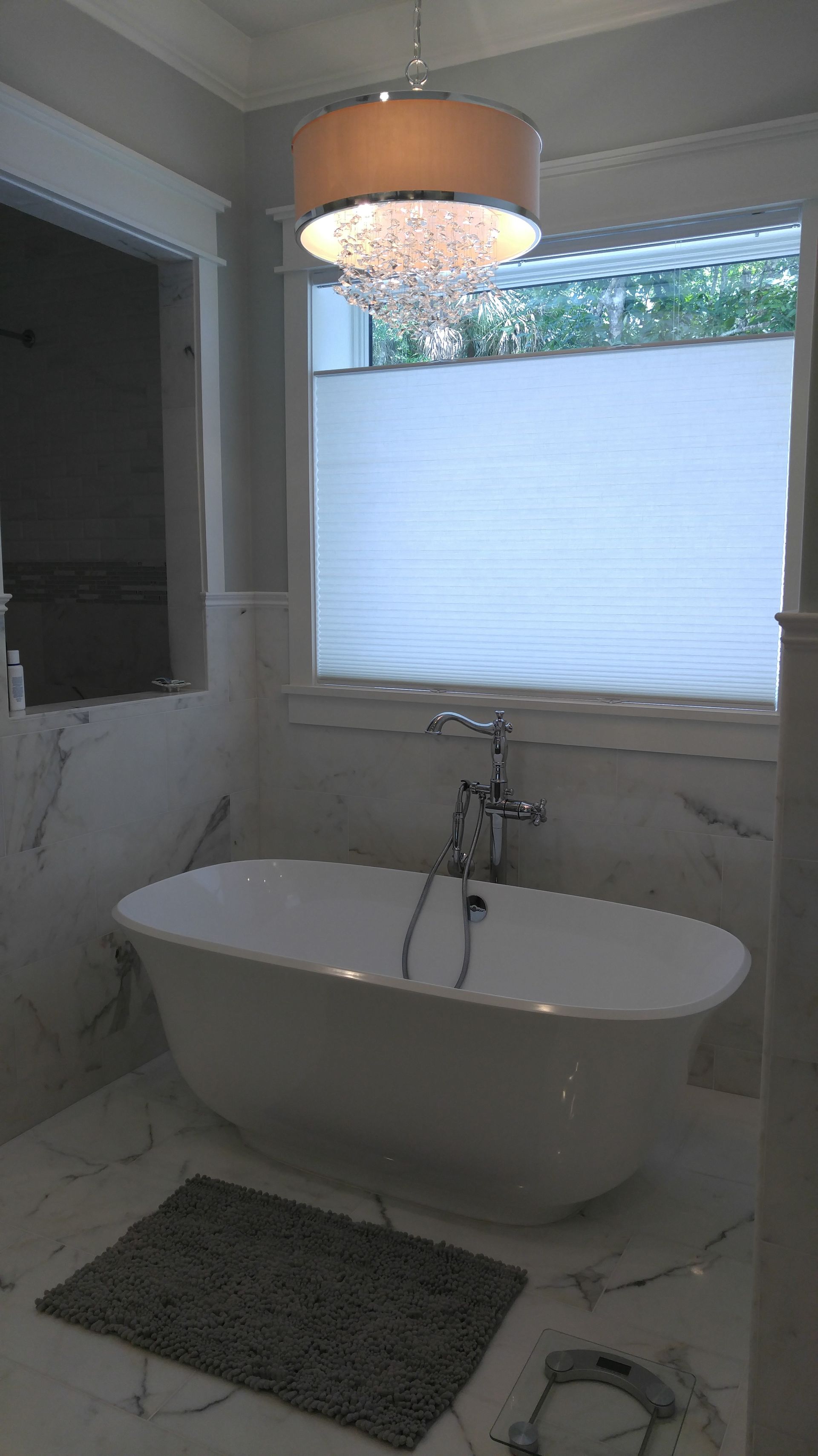 Bathroom with white tub, marble walls and floor, silver fixtures, a textured gray bath mat, and chandelier.