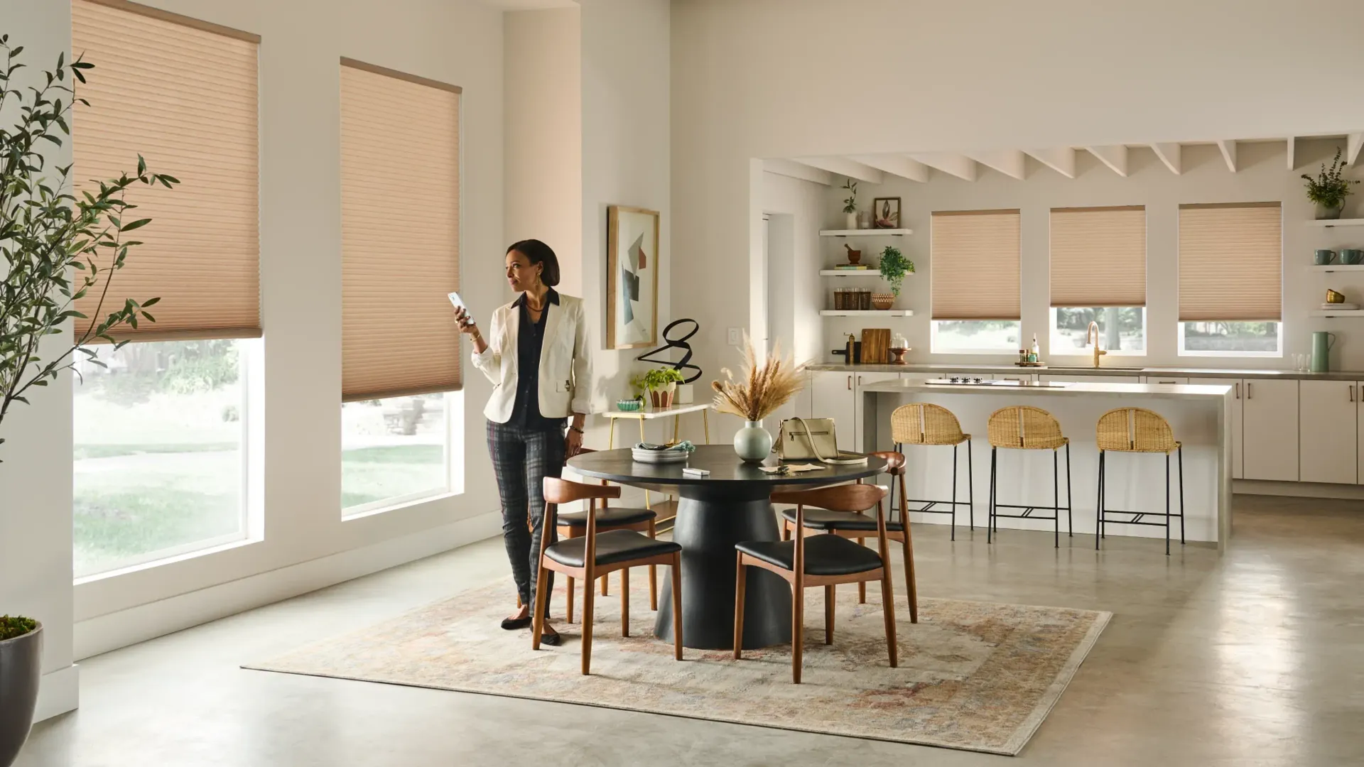 Woman controlling smart shades in dining room. Beige shades, dark table, light floor.