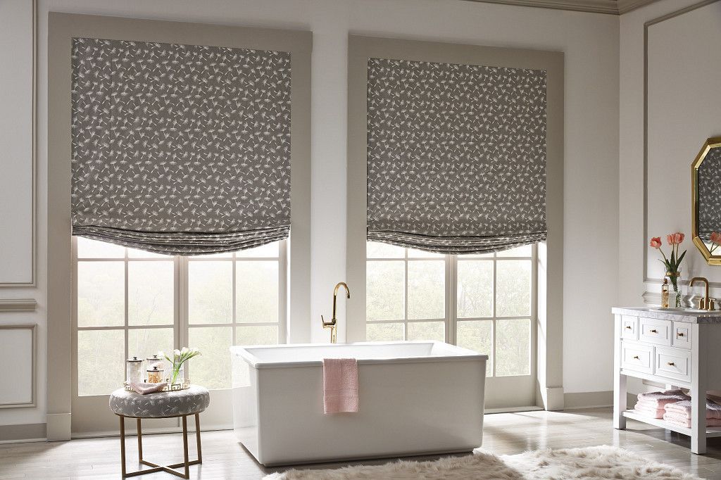Bathroom with two windows, a bathtub, and patterned Roman shades; neutral tones.