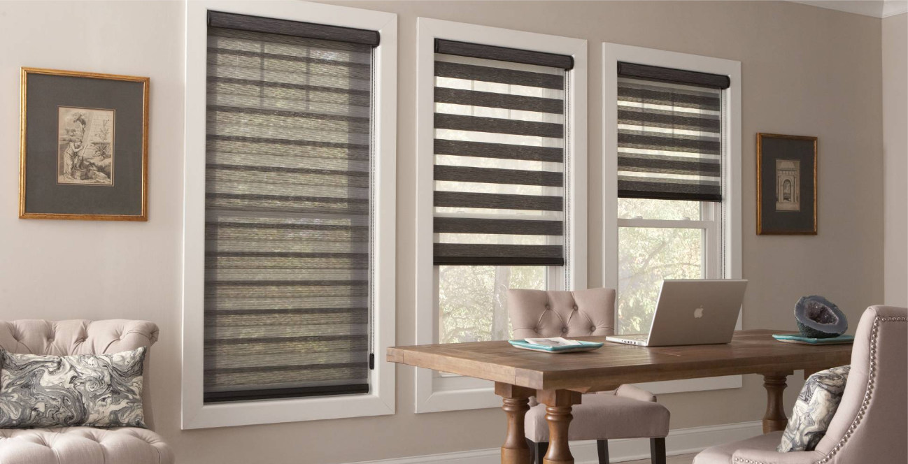 Three windows with different roller shades above a desk with a laptop; two framed pictures on the walls.