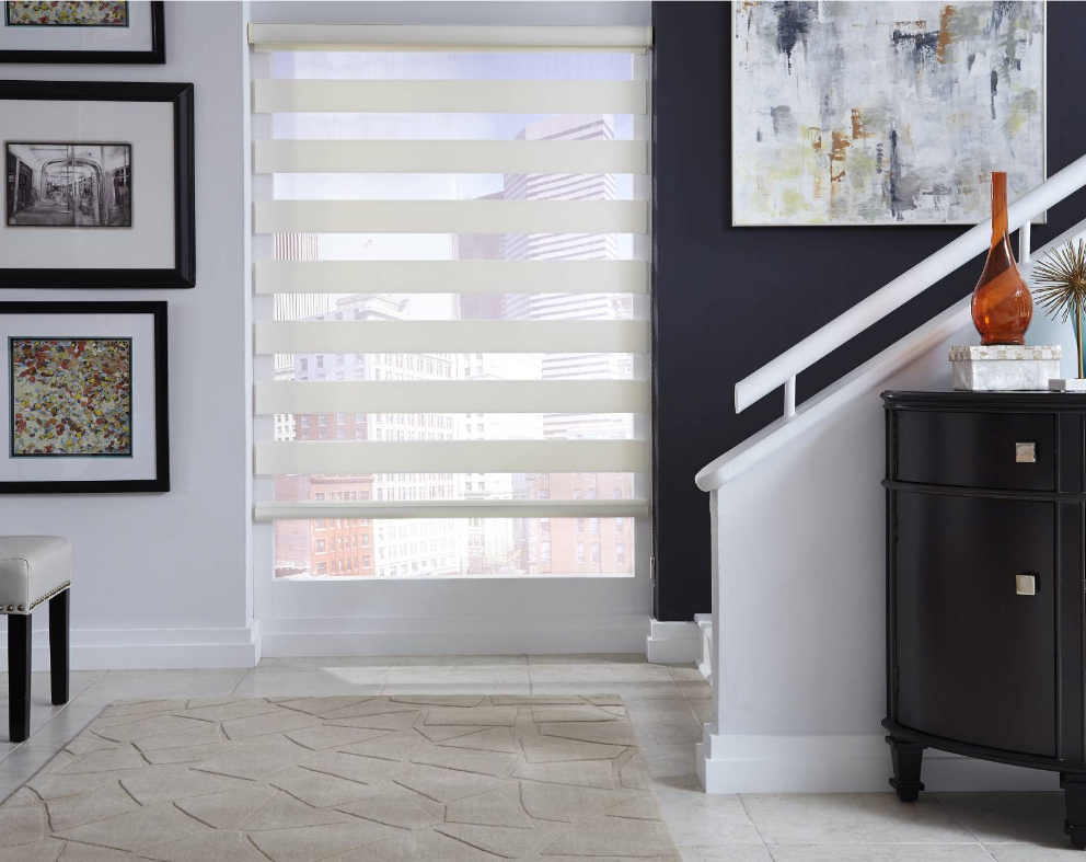 Room with layered window shade, rug, stairs, dark cabinet, art and photos on wall.