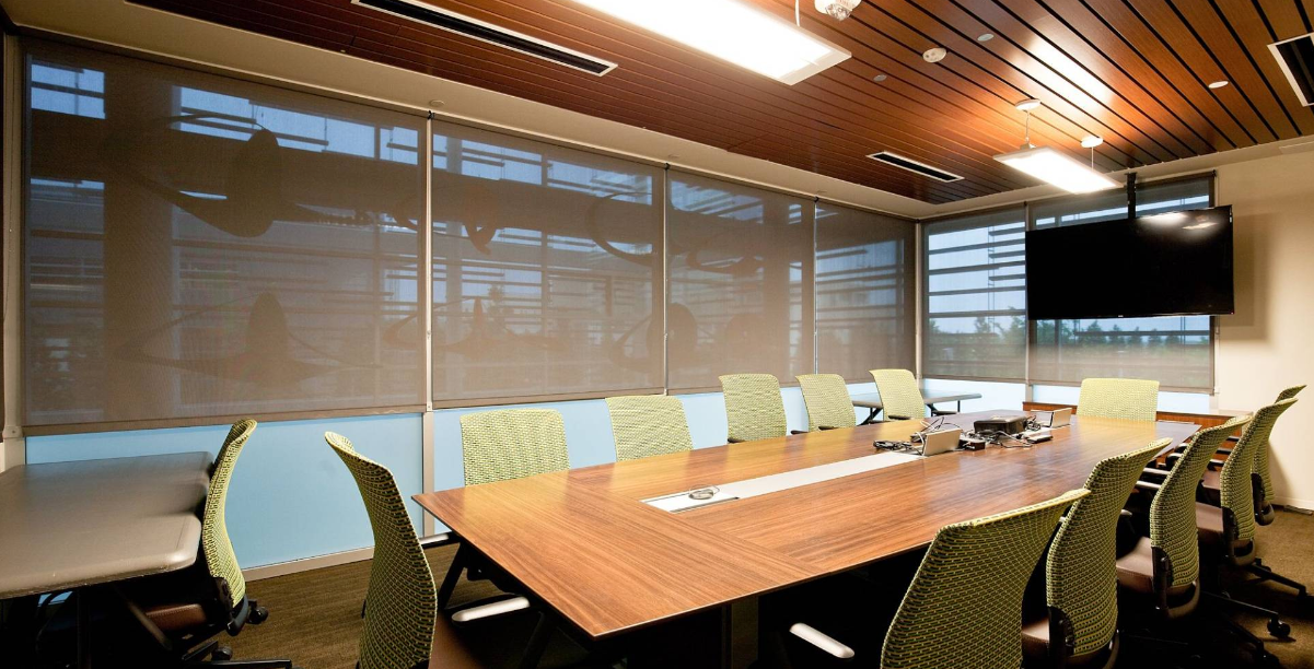 A conference room with a long table, chairs, large windows with shades, and a mounted TV.