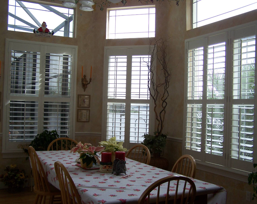 Dining room with wooden table, chairs, floral tablecloth, and large windows with white shutters.