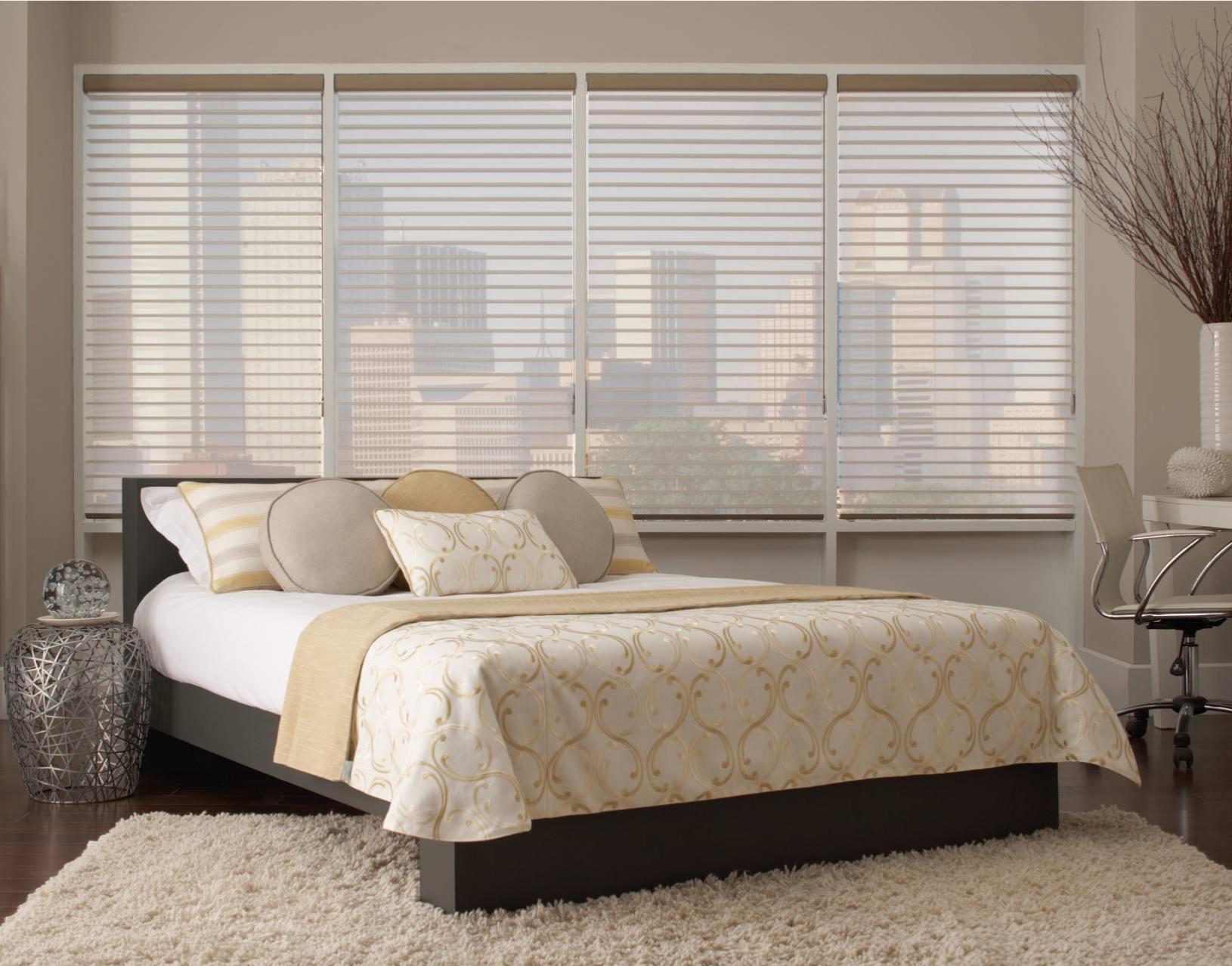 Bedroom with bed, patterned coverlet, pillows, and horizontal blinds over a window with a city view.
