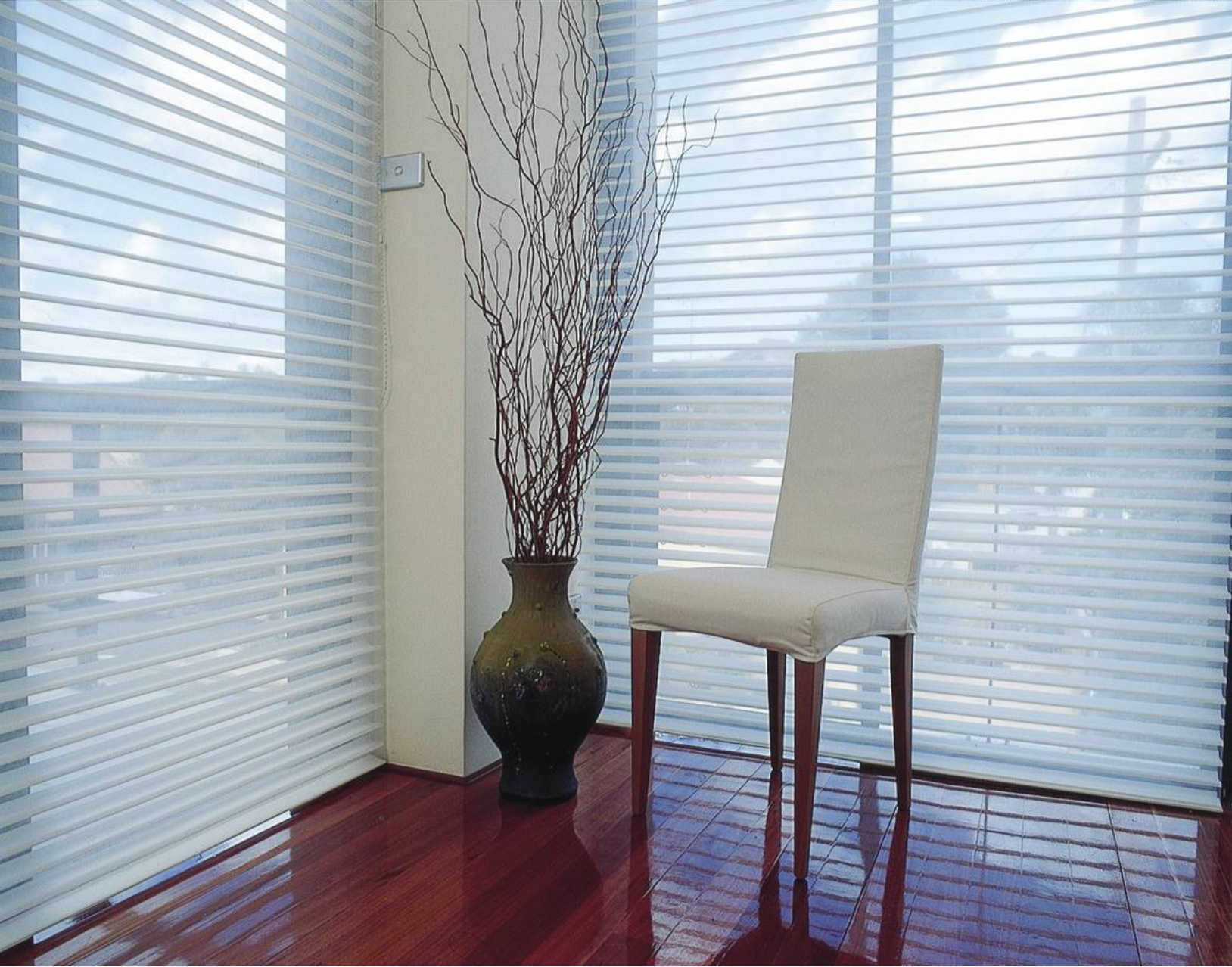 A bright room with closed white blinds, a vase of branches, and a white chair on a wood floor.