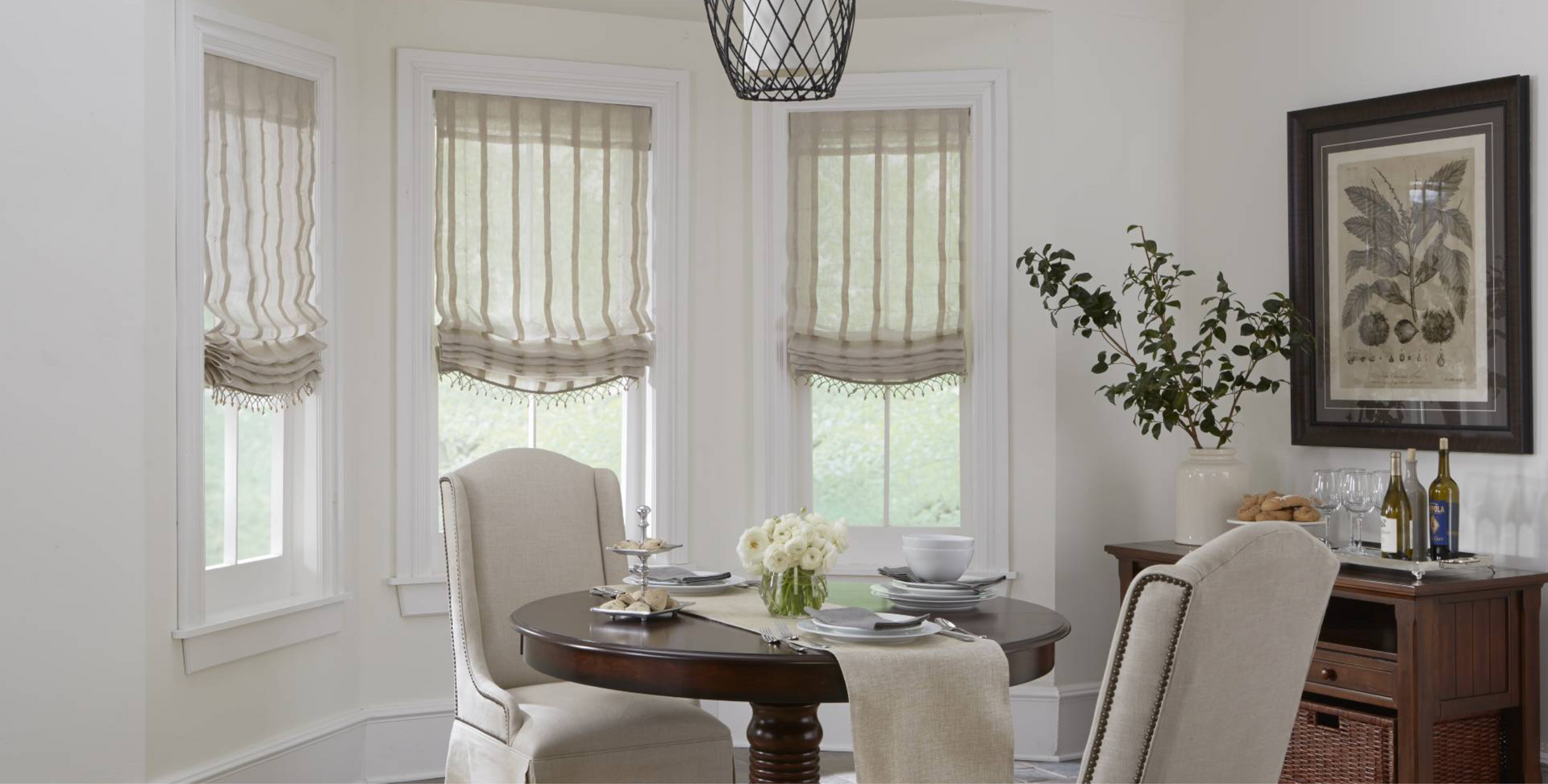 A dining room with round table, chairs, windows with Roman shades, and a sideboard.