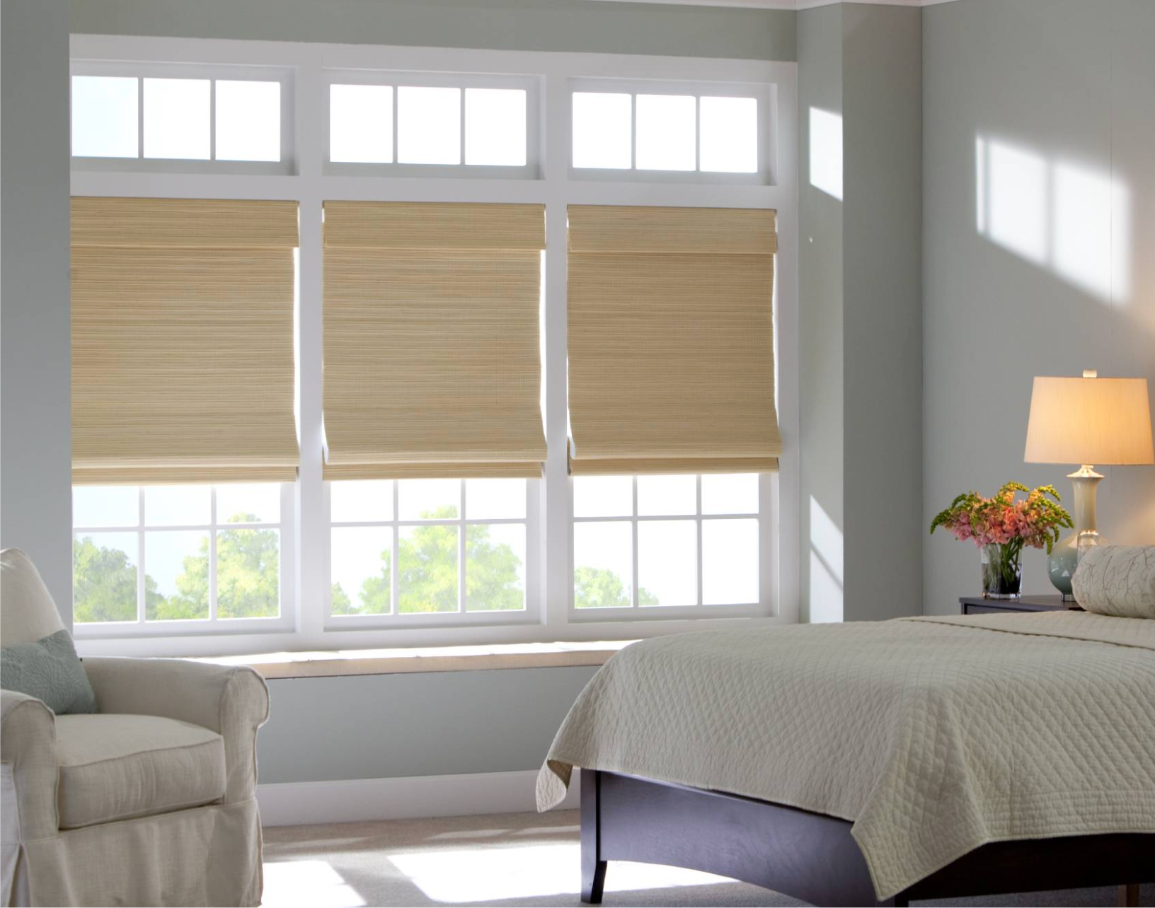 Bedroom with three windows, each with beige shades. Sunlight streams in. A bed and armchair are in view.