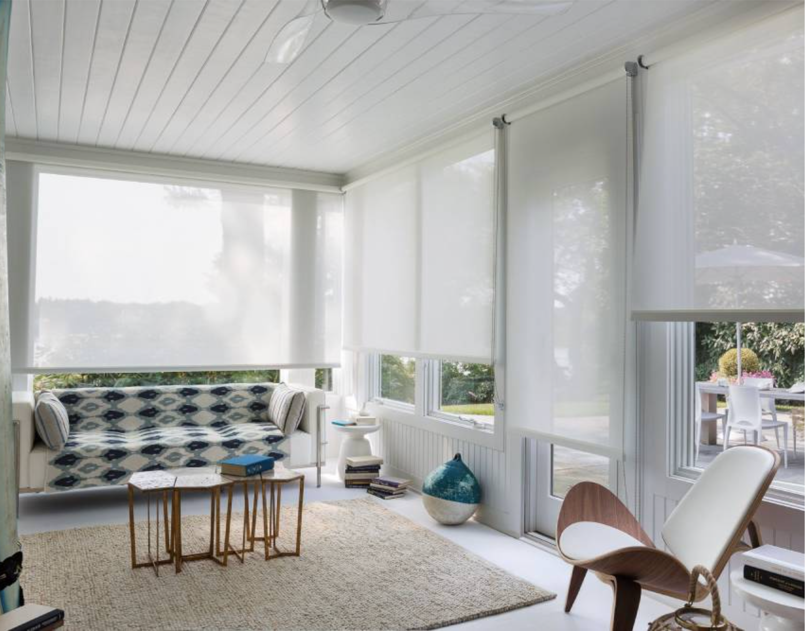 Sunroom with white roller shades, a patterned sofa, small tables, and modern chair.