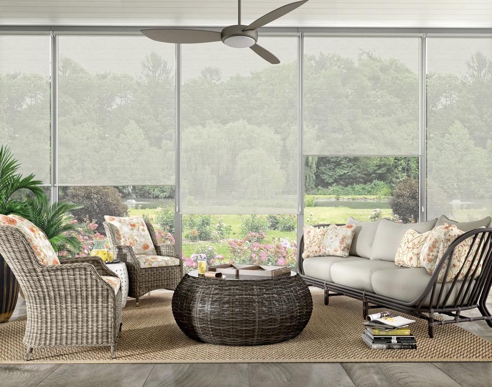 Outdoor living room with rattan furniture, a ceiling fan, and roll-up sun shades; view of a garden.