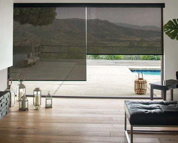 Interior with partially lowered dark roller blinds overlooking a pool and mountains. Wooden floor, modern furniture.