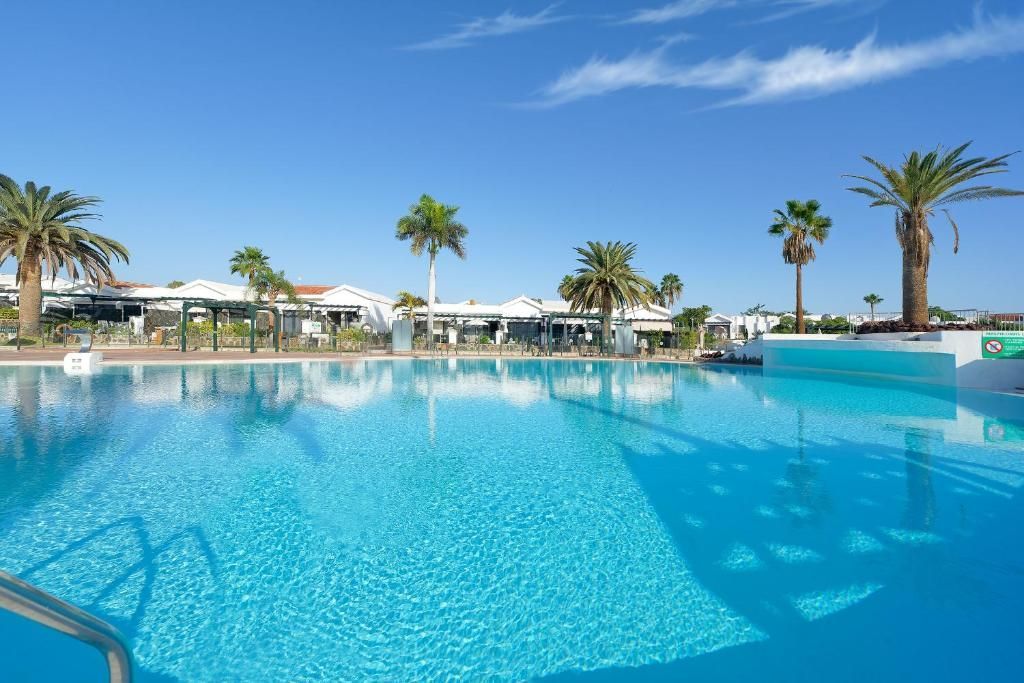 A bright blue swimming pool under a clear sky, surrounded by palm trees and white resort buildings in the background.