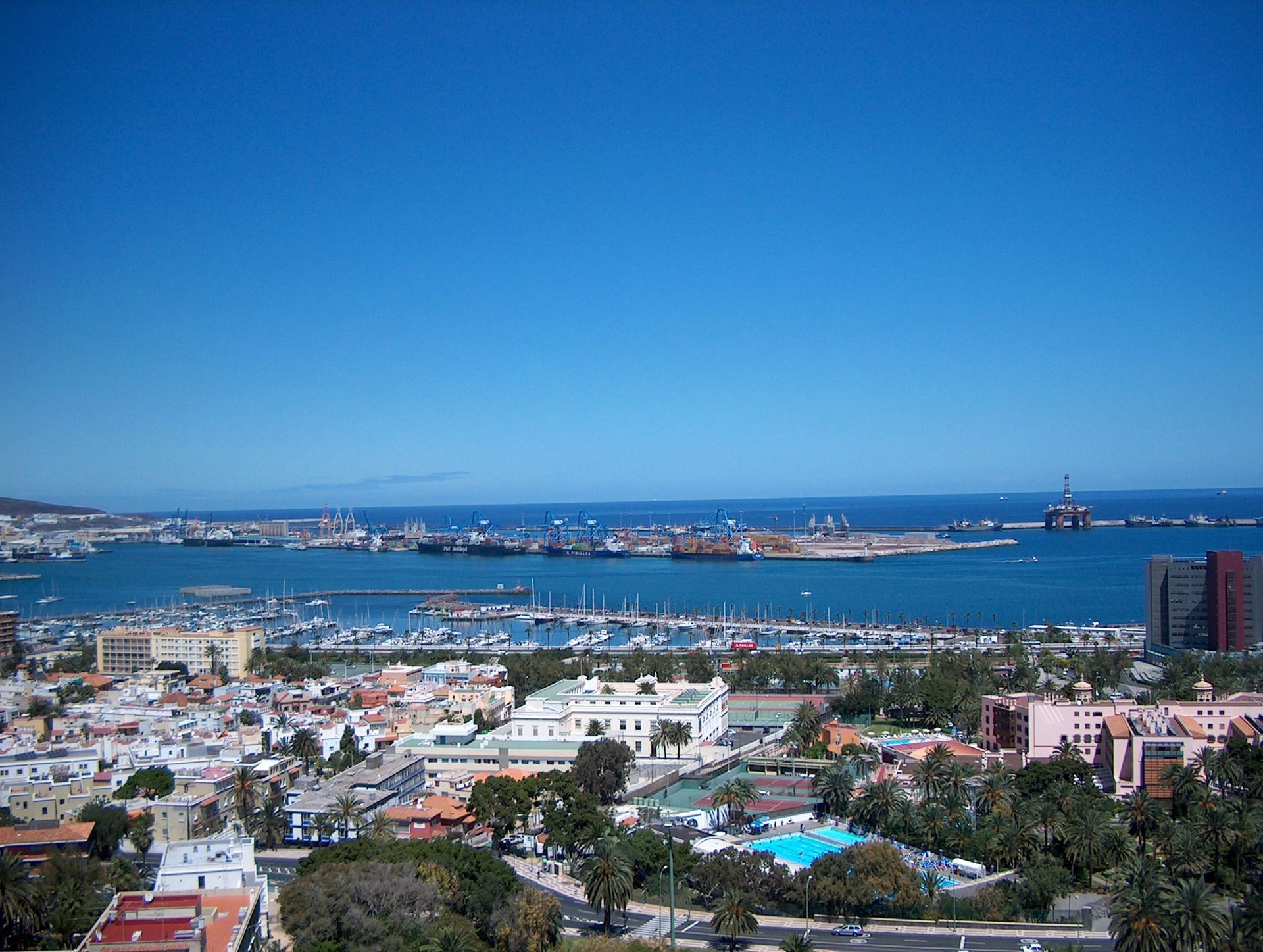 A high-angle view of a coastal city harbor with numerous boats, a large blue bay, and clear skies.