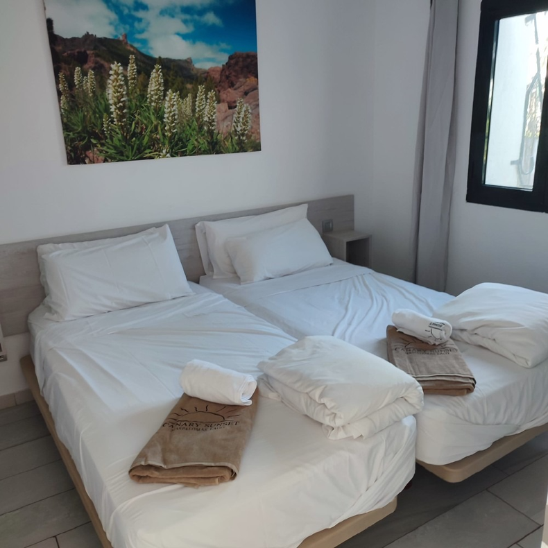 A hotel room with two white single beds, each topped with a folded white towel and tan cloth, and a nature wall art.