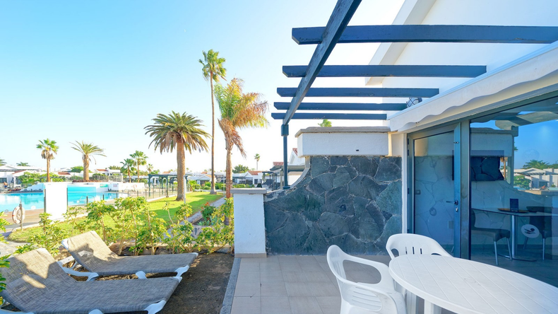 A sunny patio with lounge chairs, a table, and chairs, overlooking a resort pool and palm trees under a clear blue sky.