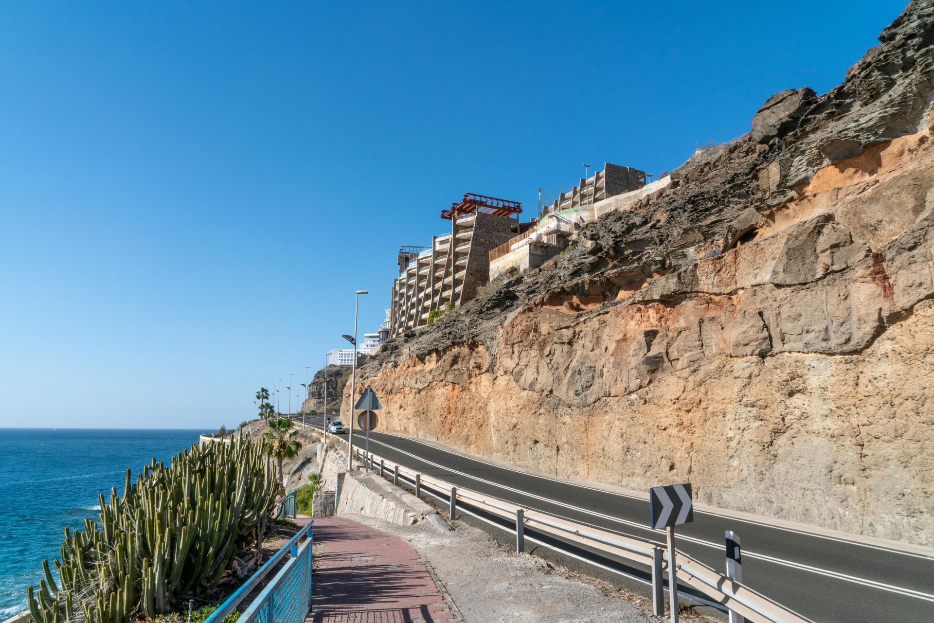 A coastal road runs alongside a rocky cliff, with buildings perched at the top under a bright blue sky.