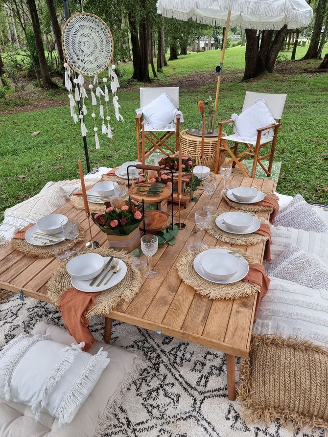 Picnic setup with a wooden table, chairs, and cushions in a forest setting.
