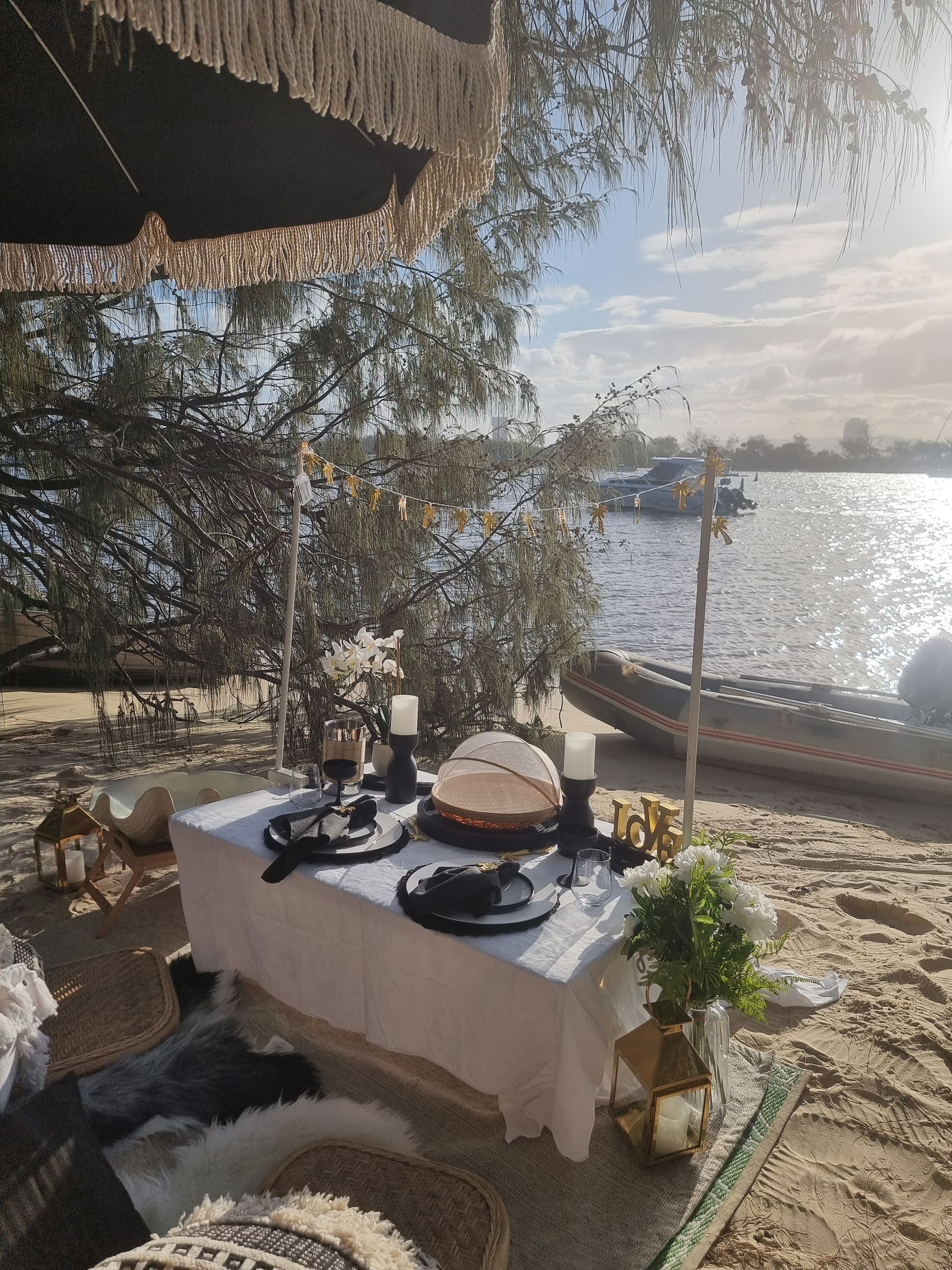 Picnic table set for a meal on a beach near water, with cake, decor, and umbrella. Sunny, outdoor setting.