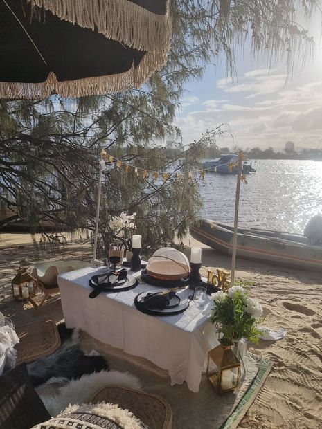 Picnic table set for a meal on a beach near water, with cake, decor, and umbrella. Sunny, outdoor setting.