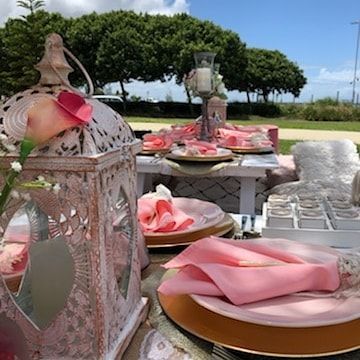 Outdoor table setting with pink napkins, gold chargers, decorative lantern, and candles.