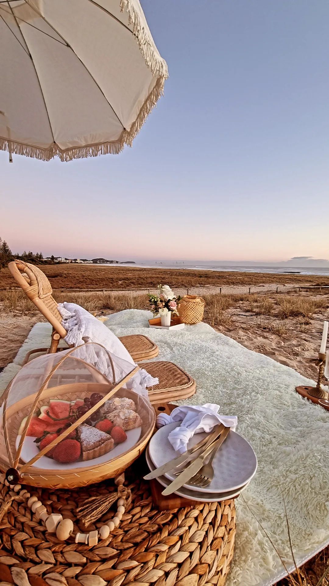 Picnic on beach with food basket, plates, blanket, and umbrella at sunset.