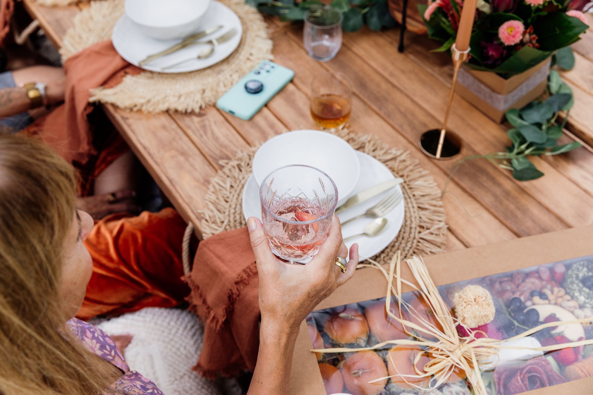Woman holding a glass, seated at a picnic table with food, drinks, and floral decorations.