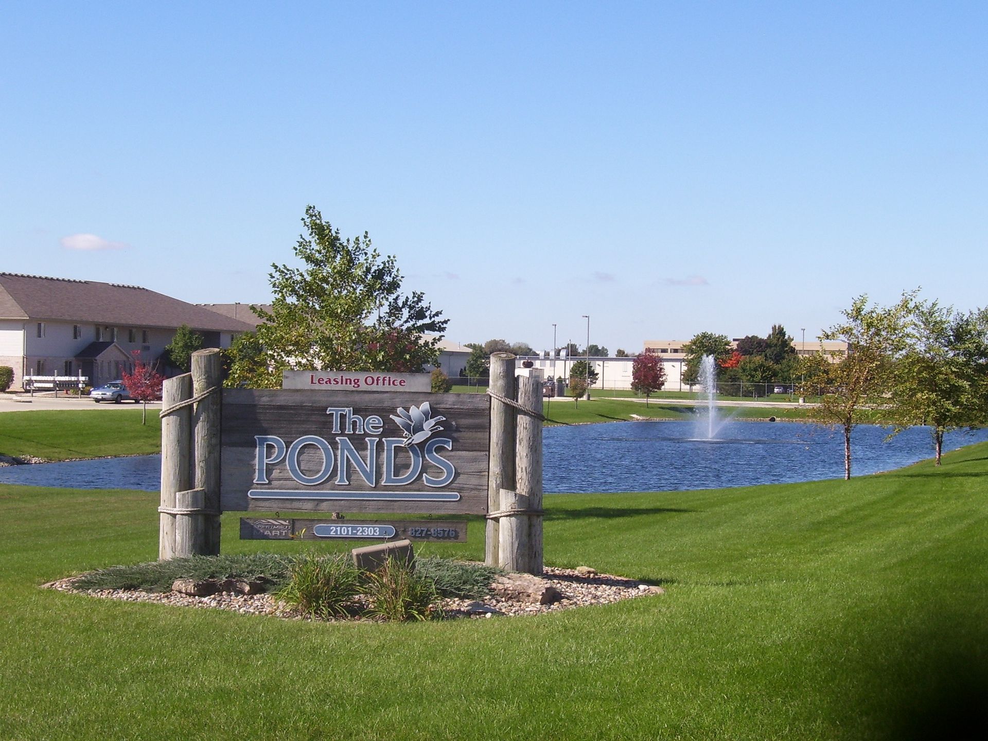 Sign for The Ponds residential area with a small pond and fountain under a blue sky.
